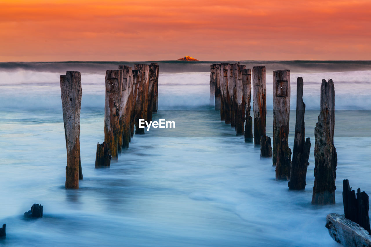 Wooden posts in sea against cloudy sky during sunset