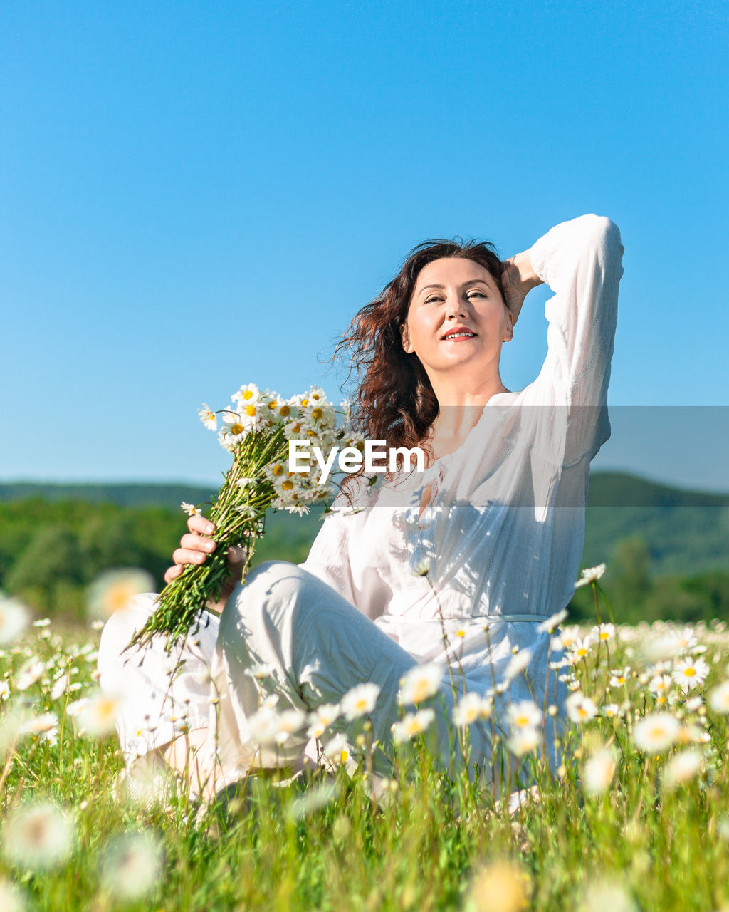 A happy woman in a spacious summer dress poses on a blooming field of daisies on a clear summer day