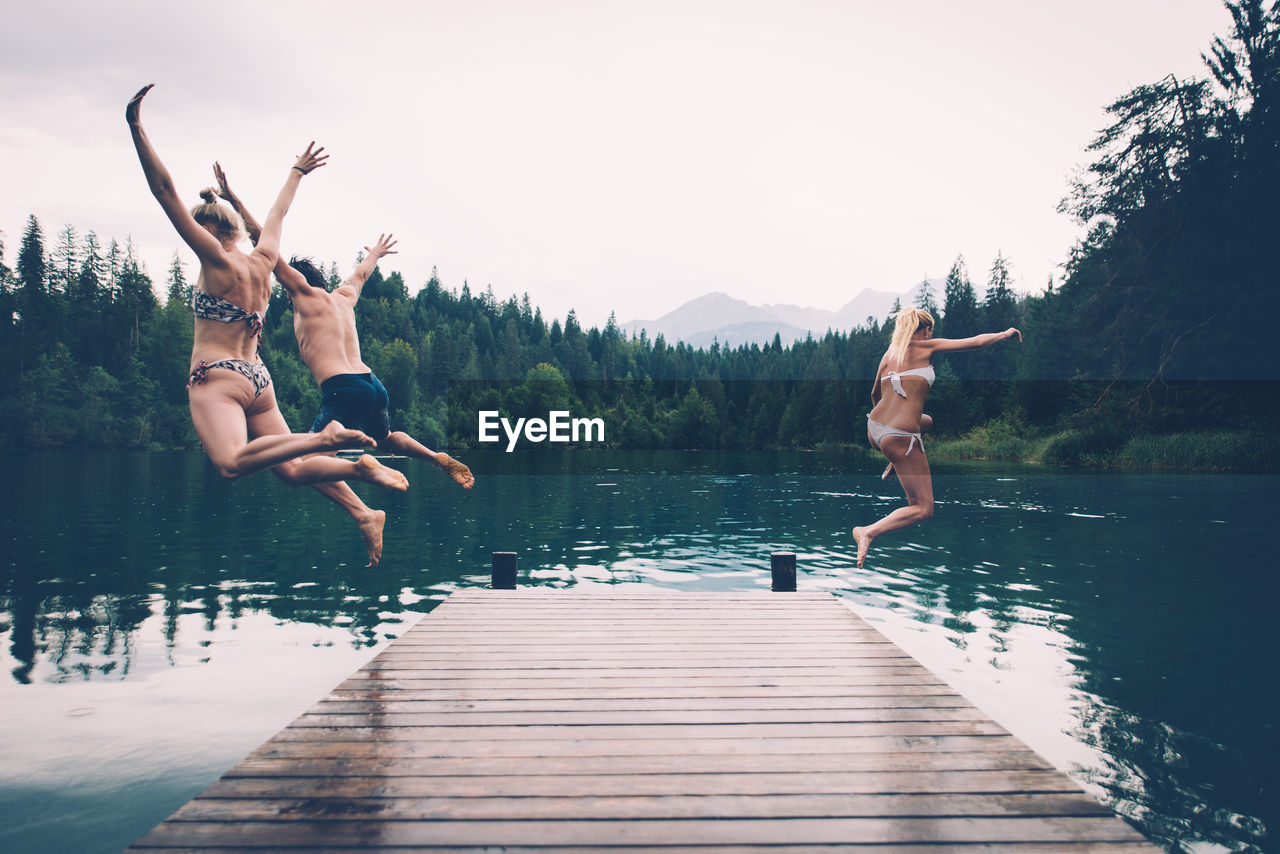 Full length of friends jumping in lake against sky