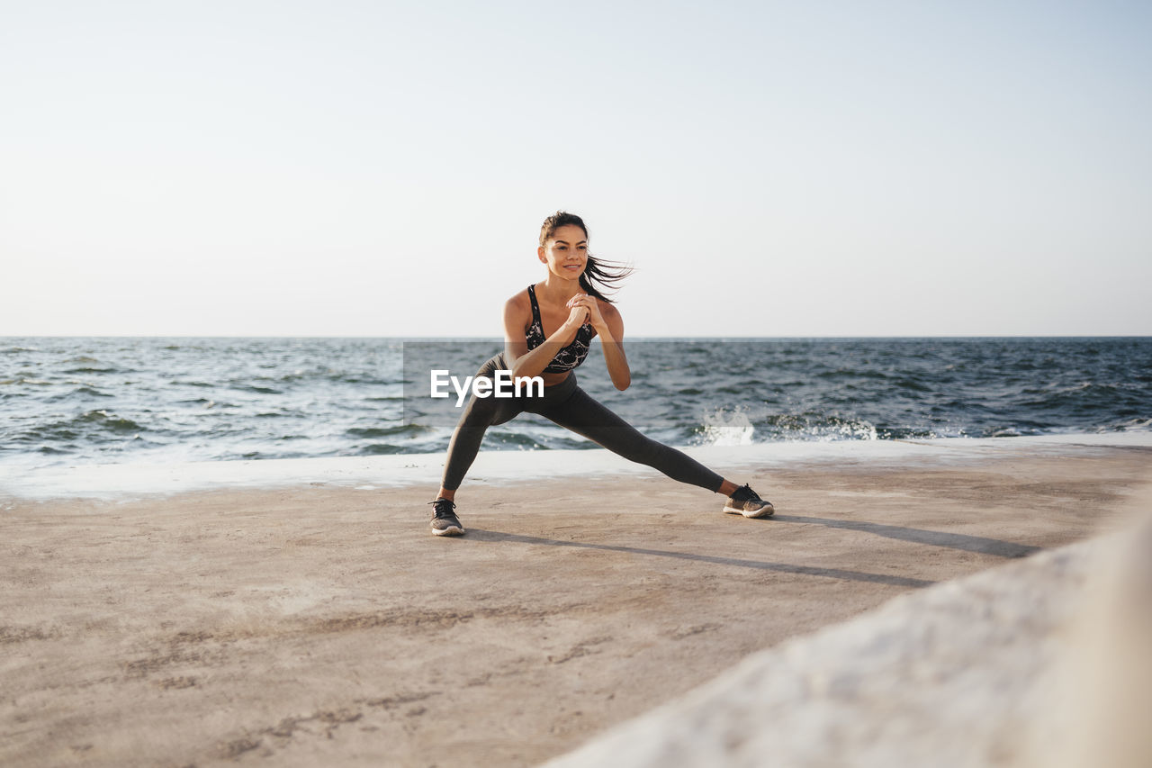 Young woman stretching legs exercising at beach on sunny day