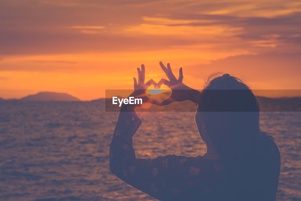 Woman making heart shape towards sun while standing by sea during sunset