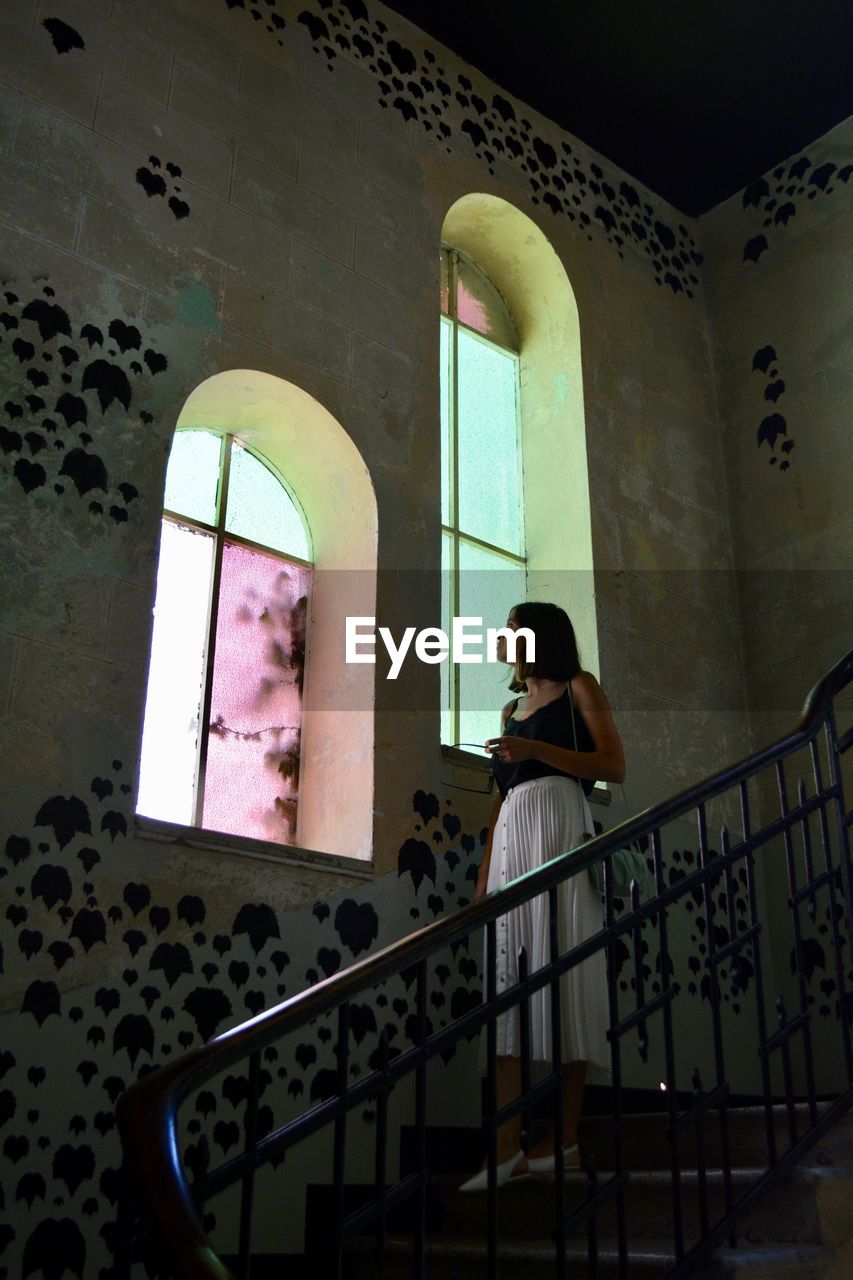 Woman looking through window while sitting on staircase in building