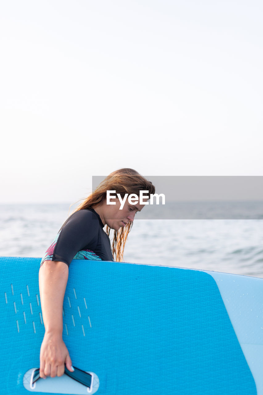 Side view of female in swimsuit standing with sup board in sea water in summer and looking away