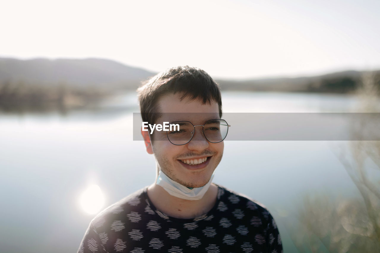 Young cheerful tourist in face mask and eyeglasses standing near river behind mountains while looking away