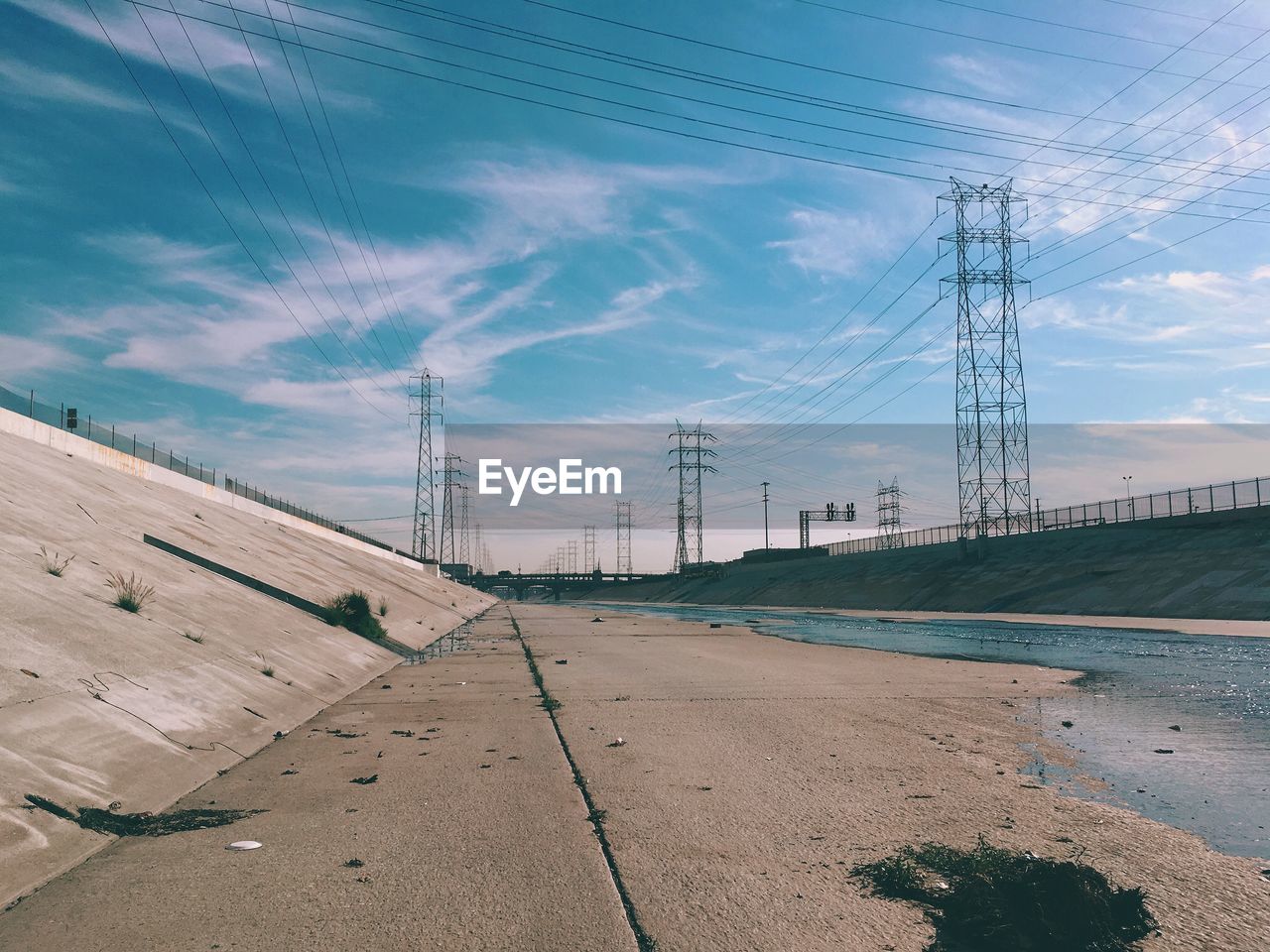 Road amidst electricity pylons against sky