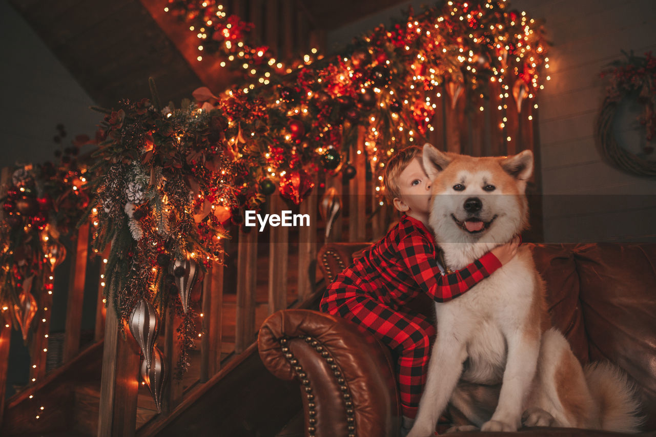 Candid authentic happy little boy in red plaid pajama hugs dog with bow tie at lodge xmas decorated