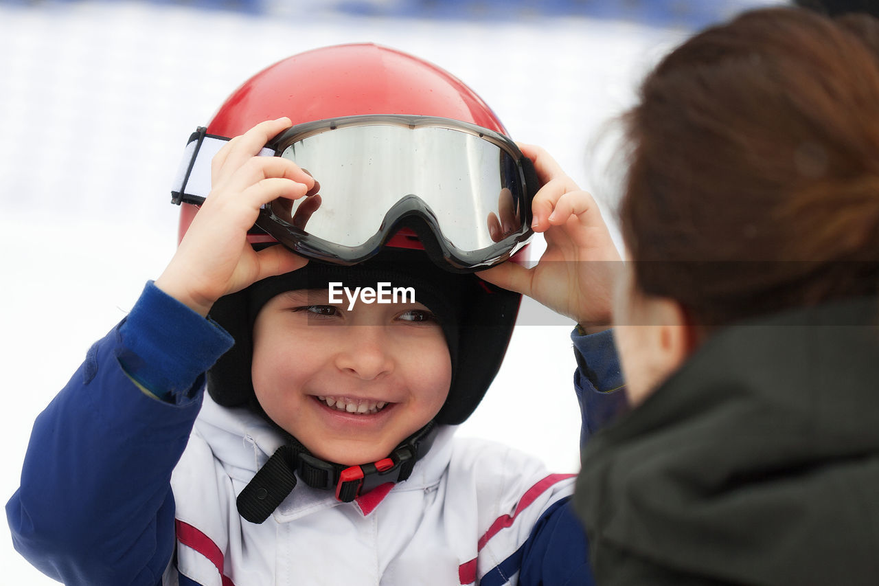 Cheerful boy in red helmet and ski goggles smiling to his mother. winter sports, young skier