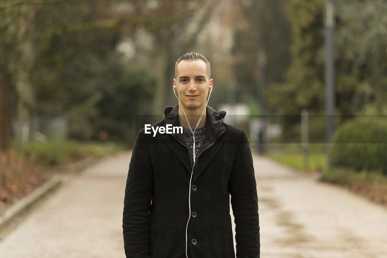 PORTRAIT OF SMILING YOUNG MAN STANDING AGAINST BLURRED BACKGROUND