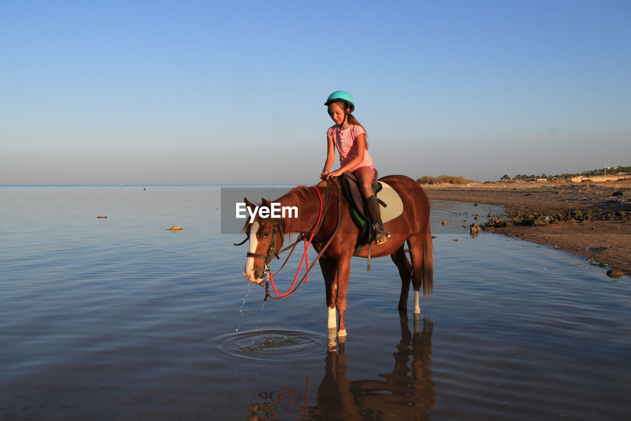 Girl riding horse in sea against blue sky