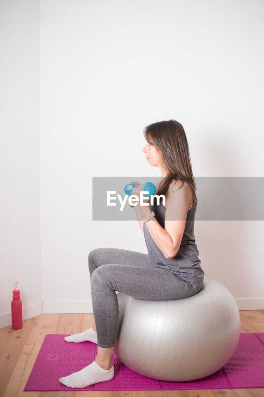 Adult caucasian woman training at home with an stability ball and dumbbell. pink yoga mat