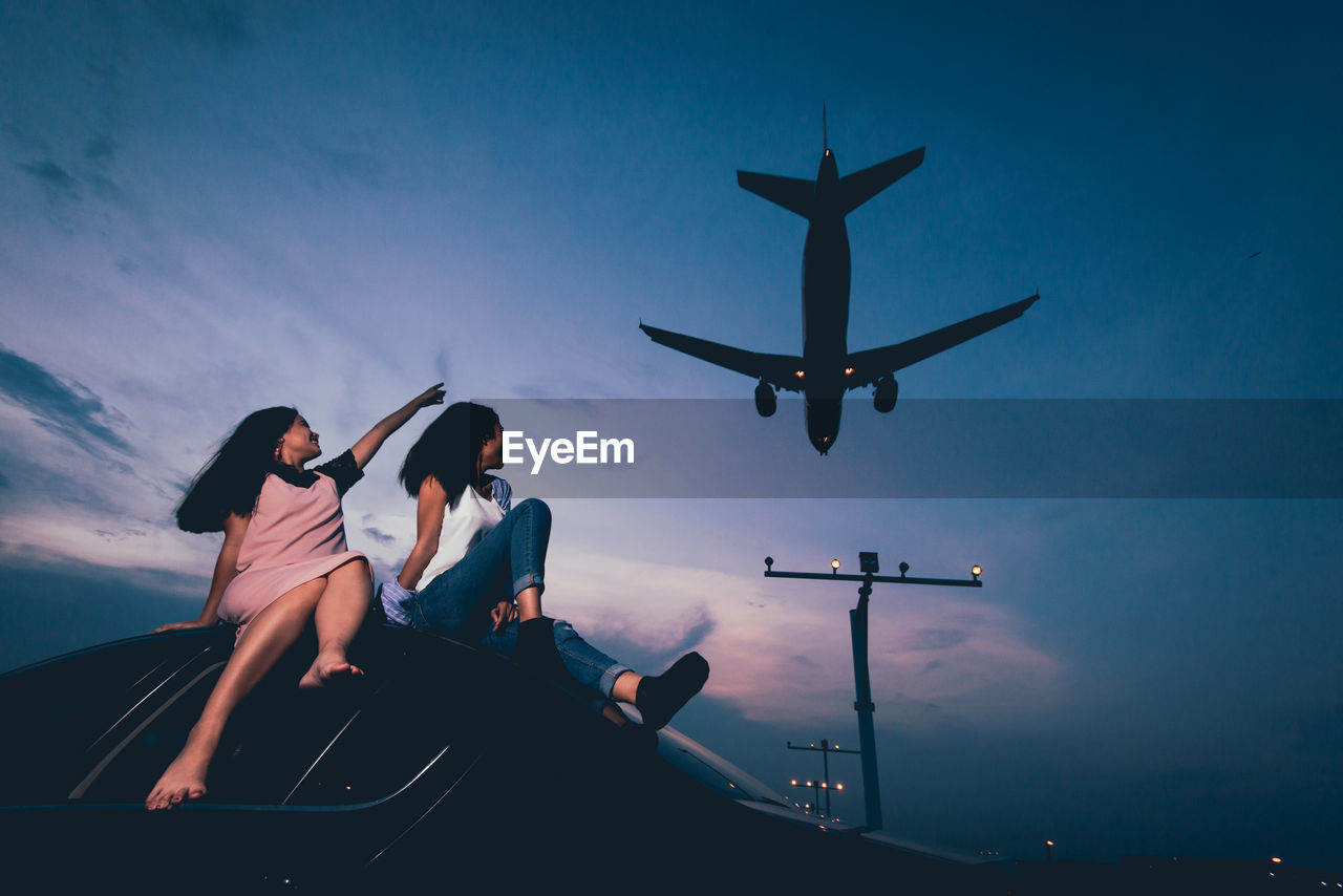 Smiling woman sitting on car with airplane landing at airport against blue sky