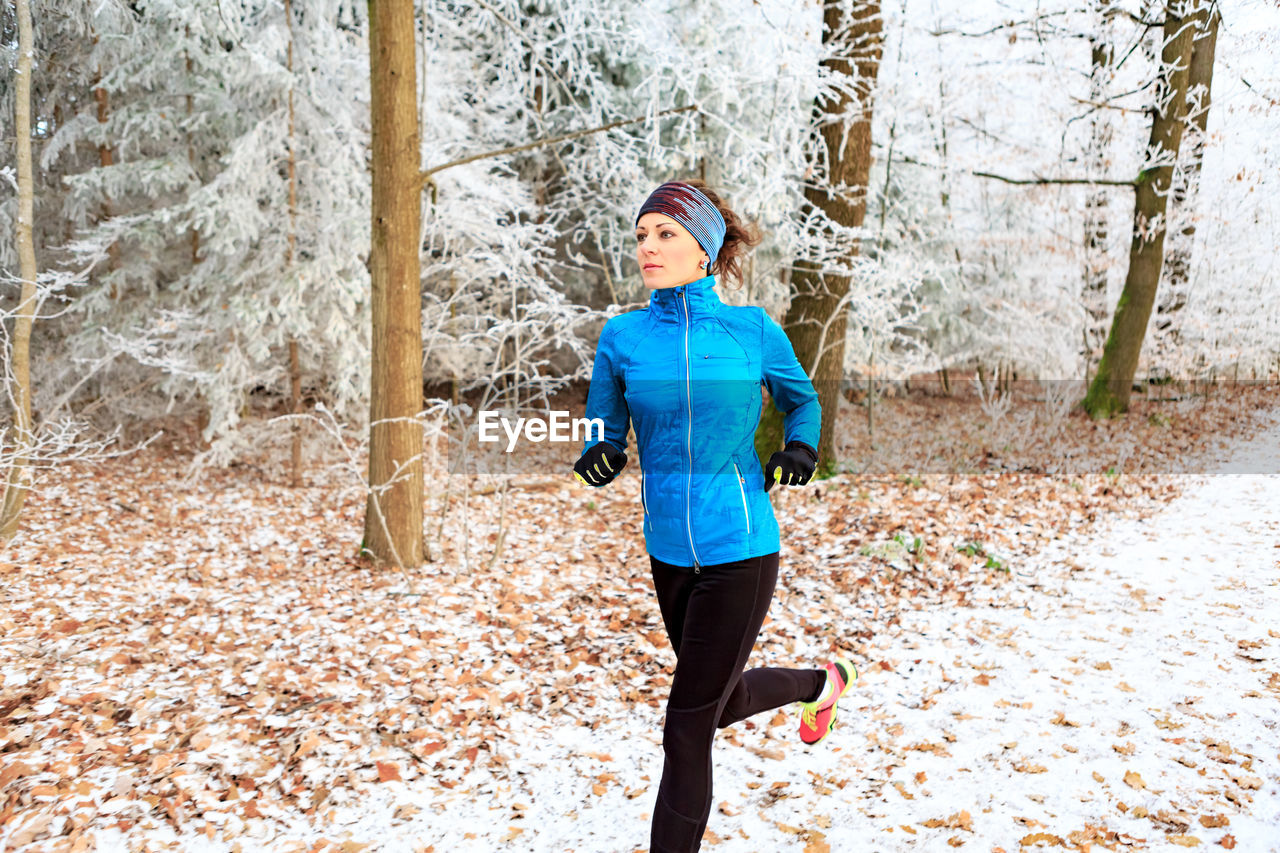 Young woman running on snow covered road in forest