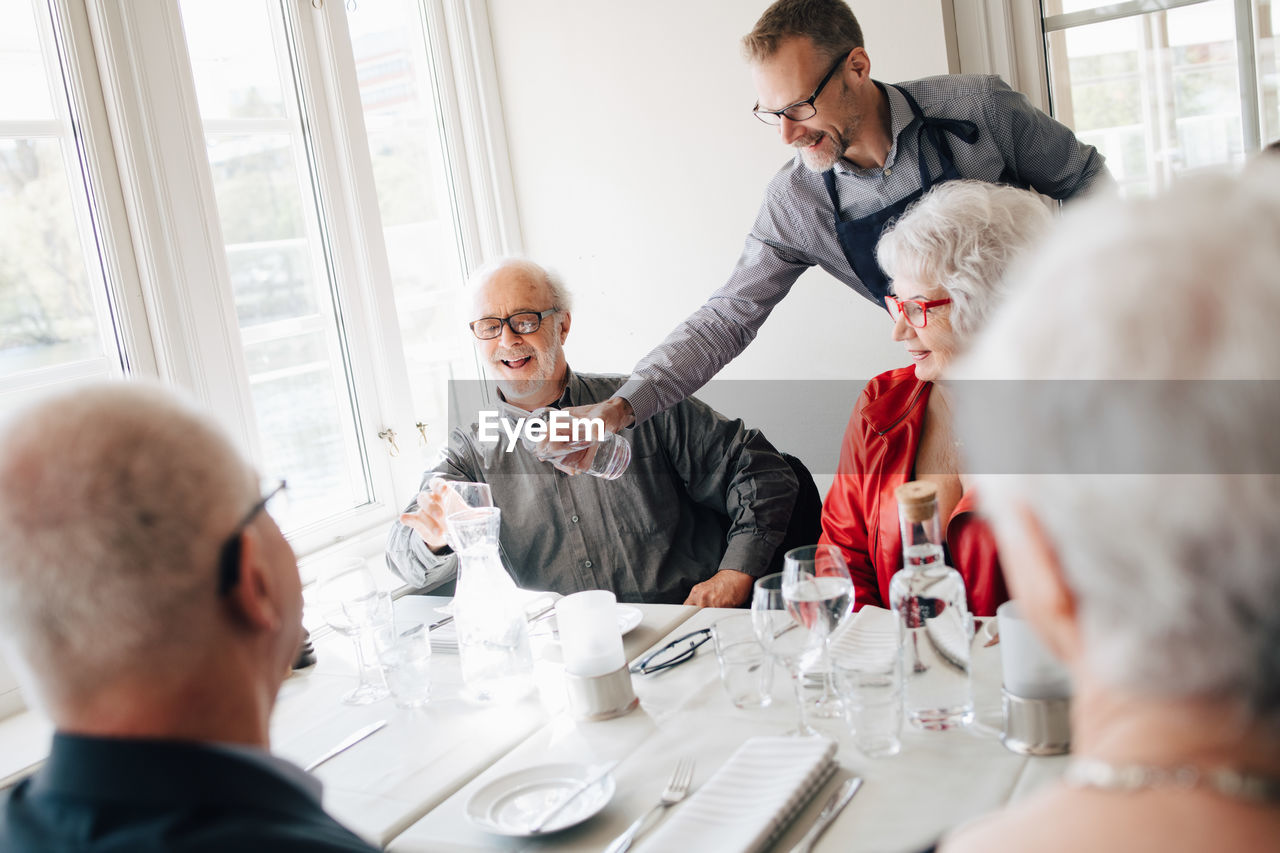 Owner serving water to smiling senior man sitting with friends in restaurant