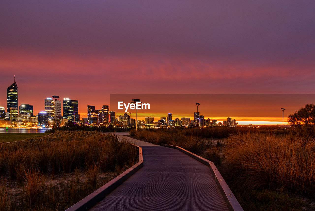 Road by illuminated buildings against sky during sunset