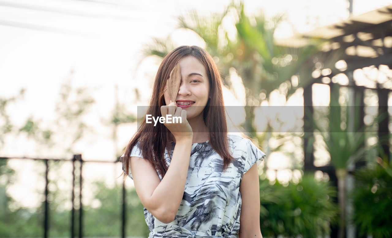 Portrait of smiling young woman covering eye with leaf