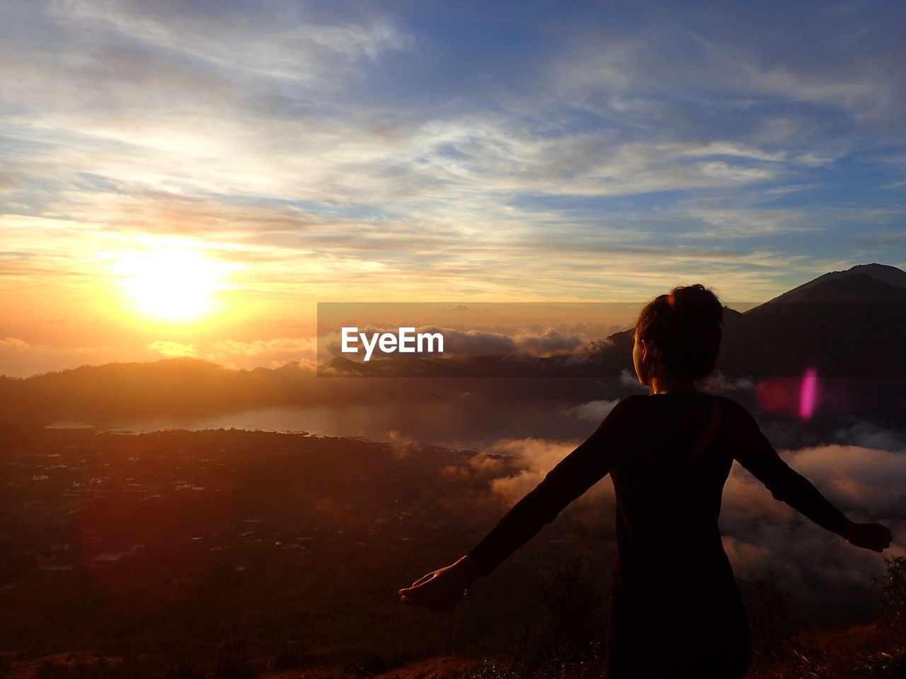 Rear view of silhouette woman with arms outstretched standing on mountain against sky during sunset