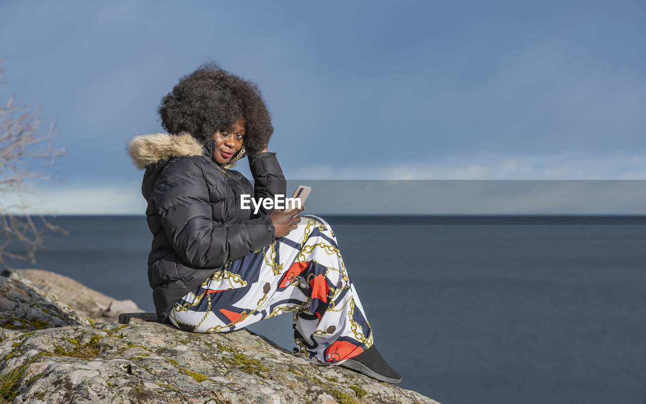 Woman with big hair sits on the rocks with a mobile phone in her hand during the spring sunset