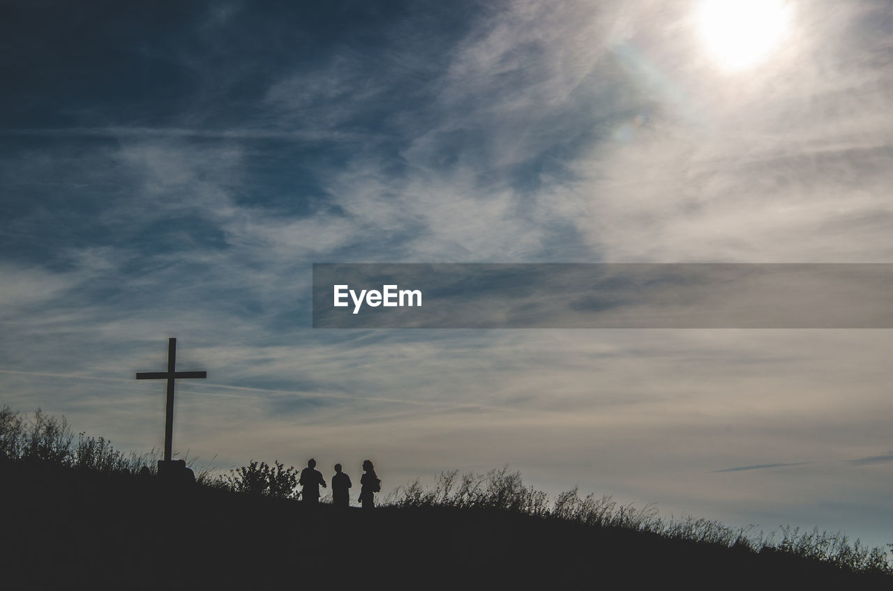 Silhouette people standing by cross on field against sky during sunset