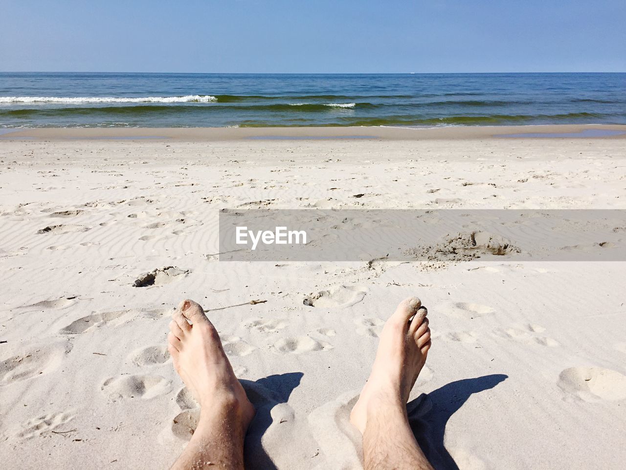 Low section of man relaxing on sand at beach against clear blue sky