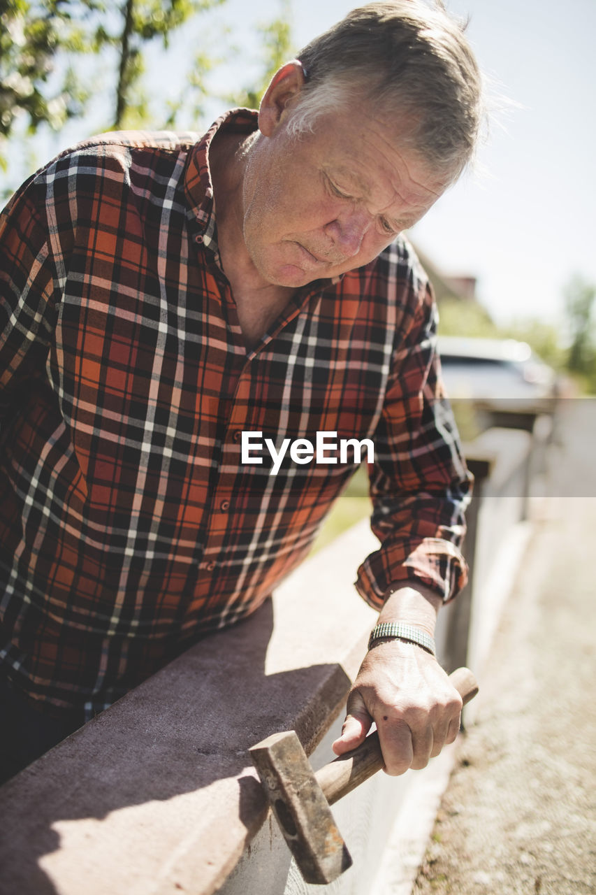 Senior man examining surrounding wall with hammer at yard