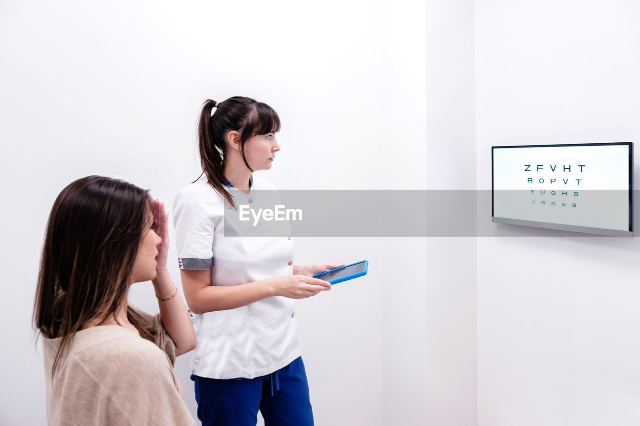 Female optometrist examining eyesight of female patient with digital test chart during visit in modern clinic