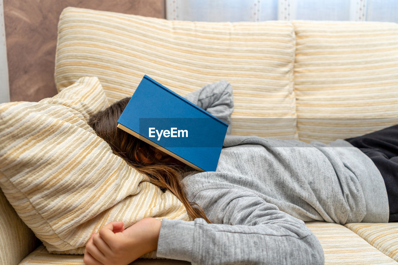 Girl with book on face relaxing over sofa at home