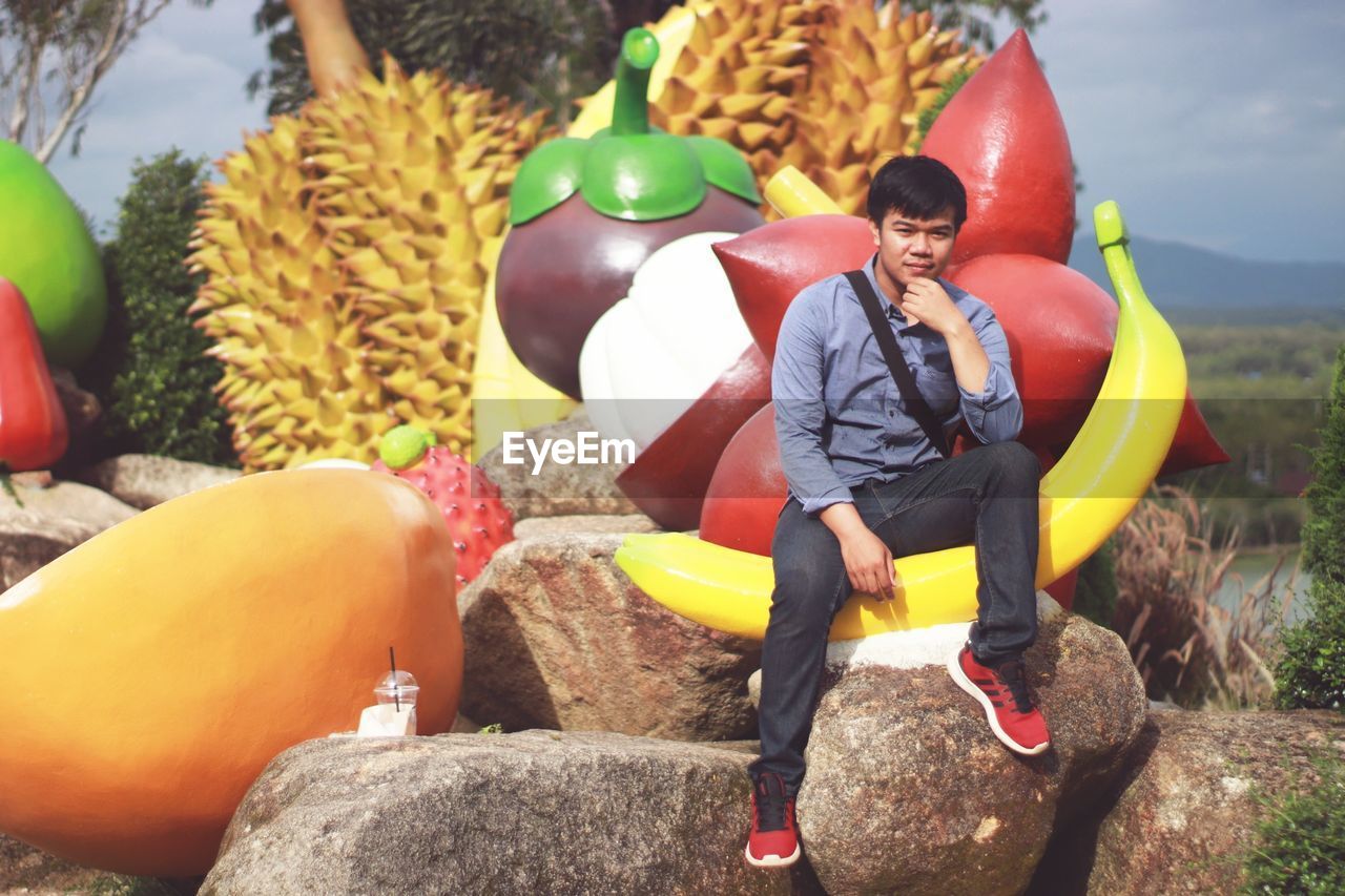 Full length portrait of man sitting on rock against artificial fruits