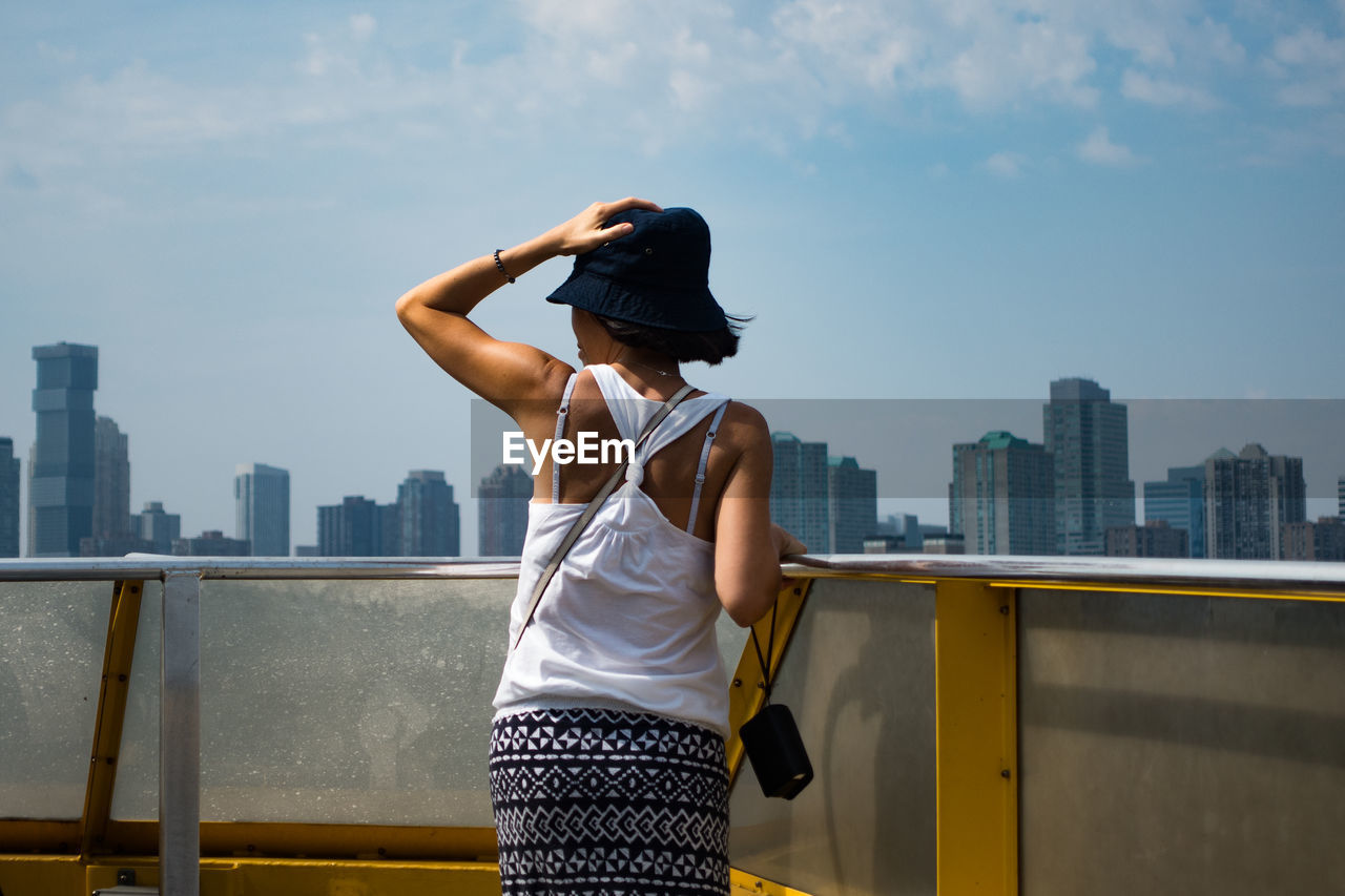 Rear view of woman holding hat while standing by railing against sky