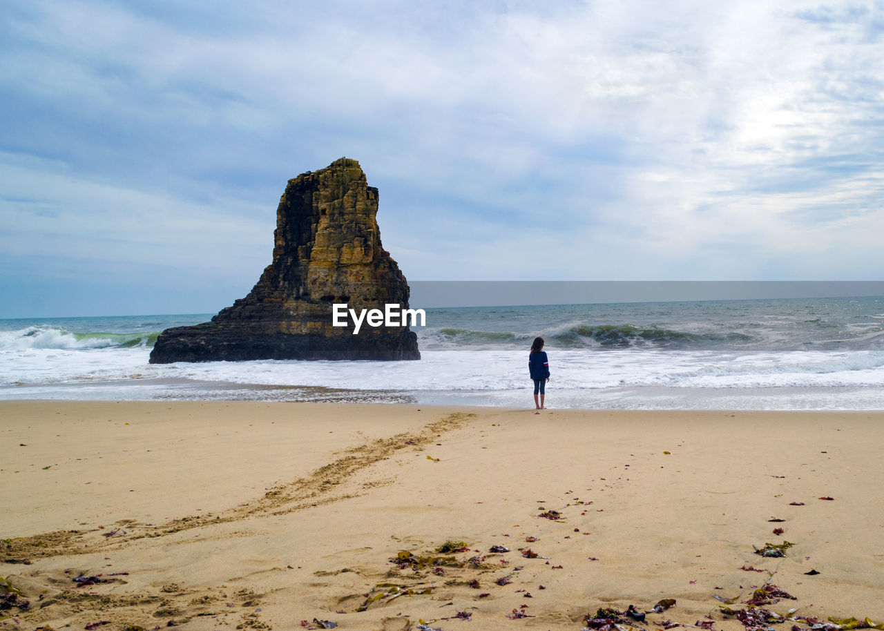 Rear view of person on beach and rock outcropping monolith against sky