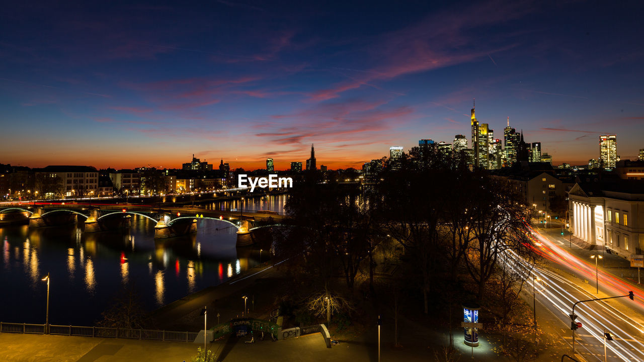 HIGH ANGLE VIEW OF ILLUMINATED BRIDGE OVER RIVER AMIDST BUILDINGS IN CITY