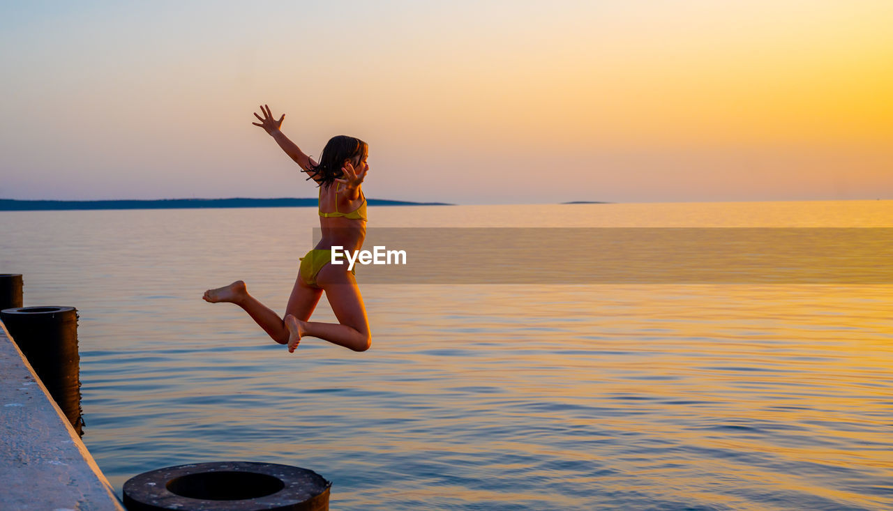 Girl with arms outstretched jumping in sea against sky during sunset