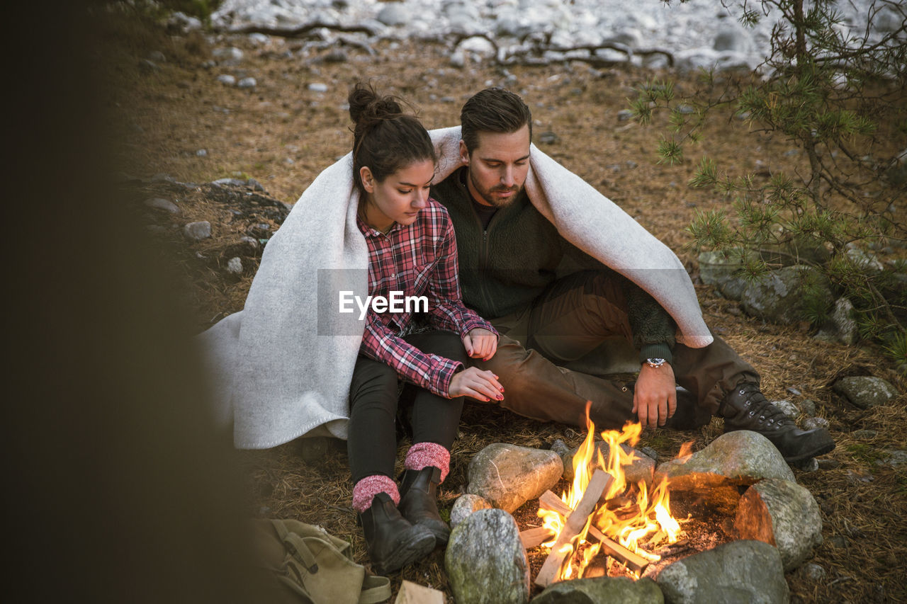 Couple wrapped in blanket while sitting by fire pit at campsite