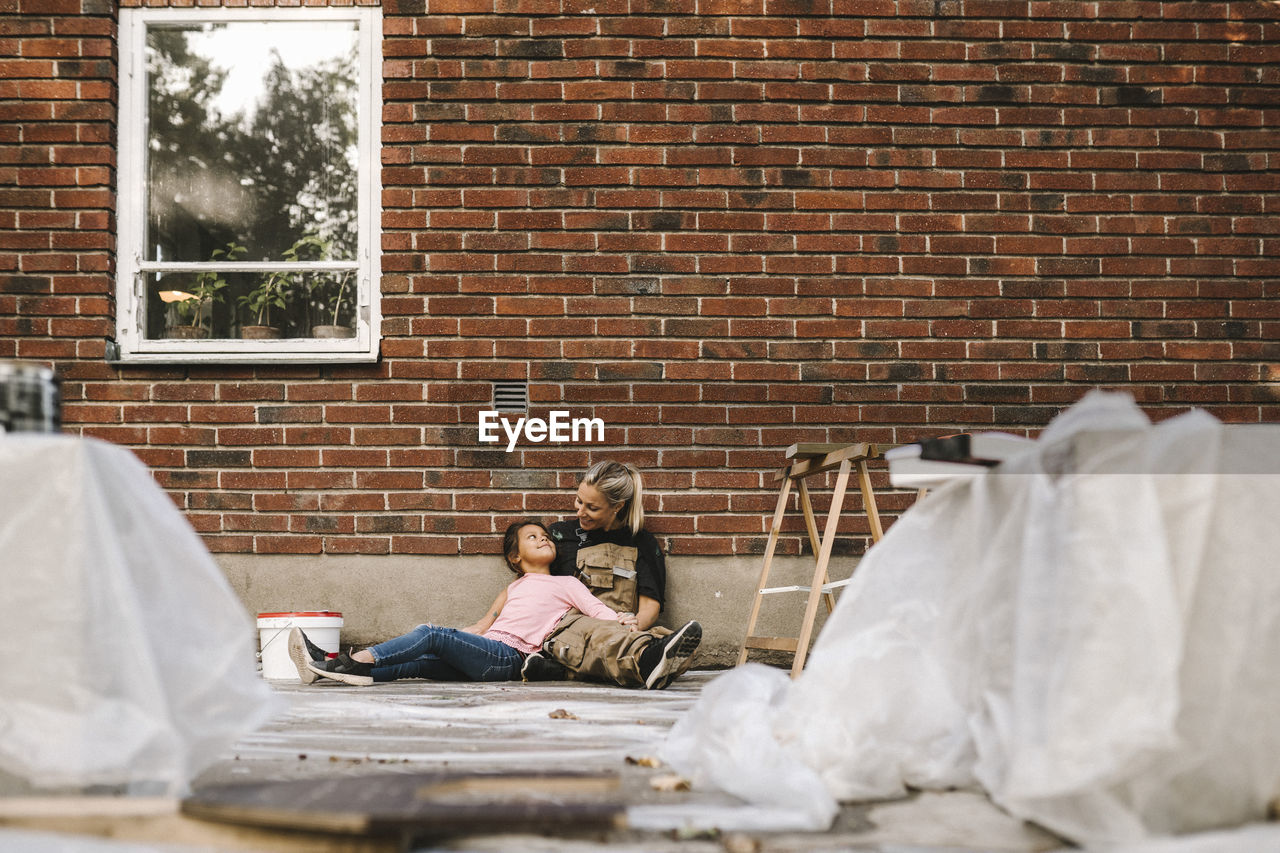 Smiling mother and daughter talking while sitting against brick wall