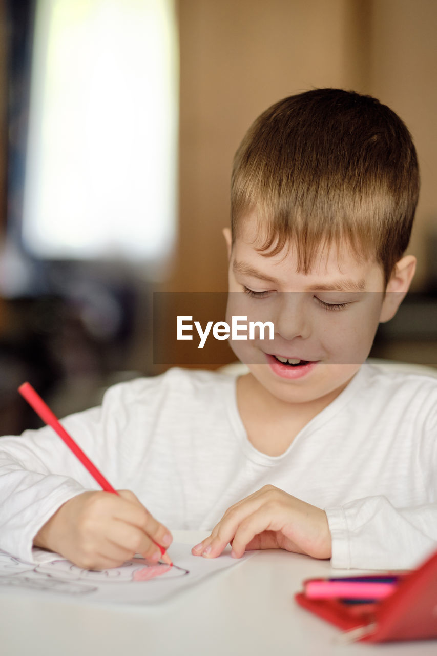 Cute and smiling cucasian boy draws with colored pencils while sitting at the table. 