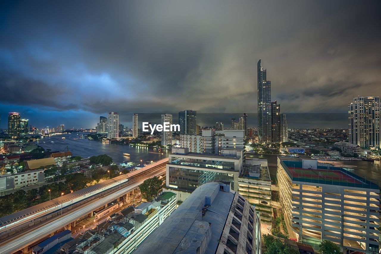 Modern illuminated buildings and chao phraya river against cloudy sky at dusk