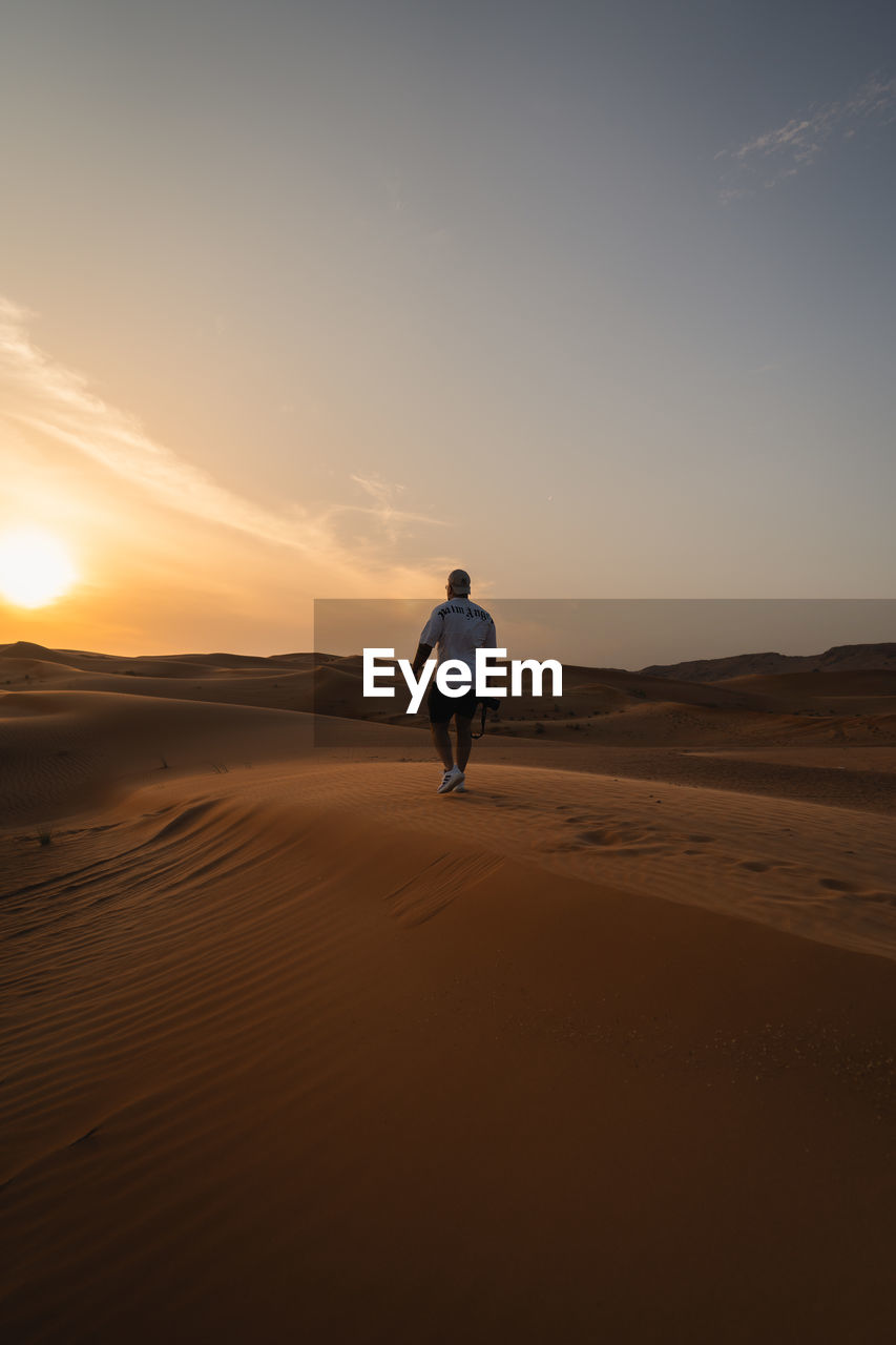 Rear view of man standing at desert against sky during sunset