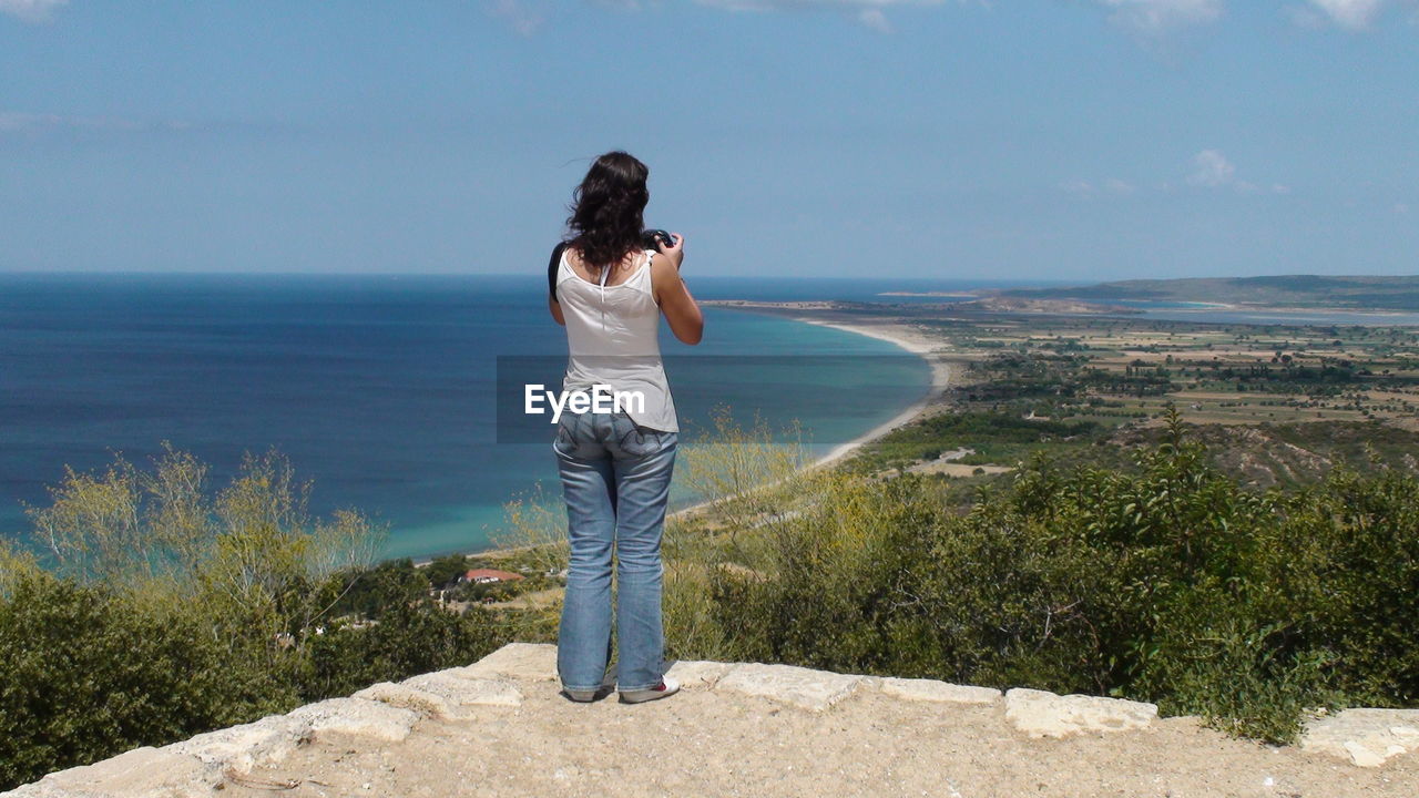 Rear view of woman photographing coastline against sky