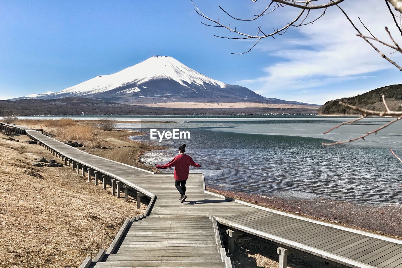 Rear view of woman walking on boardwalk at lakeshore against snowcapped mt fuji