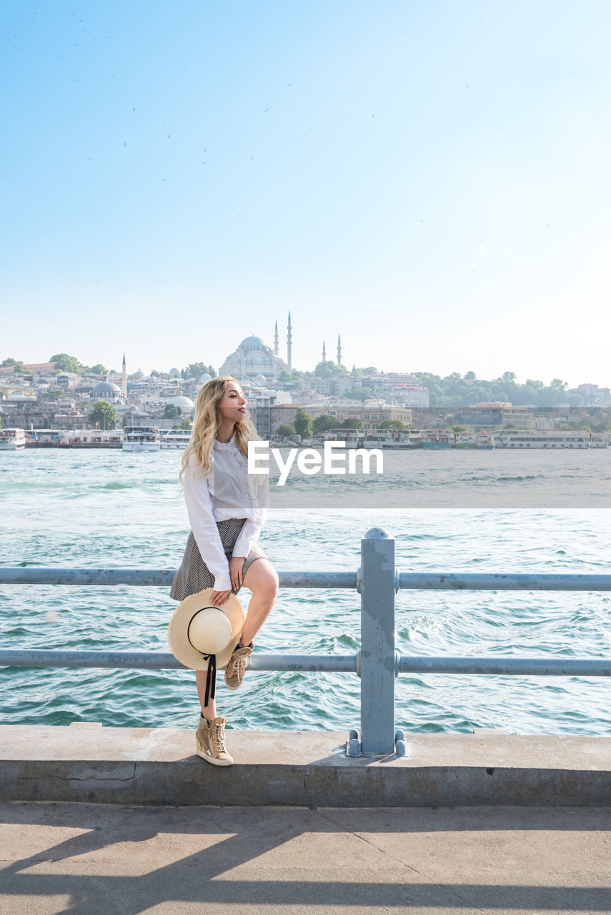 Beautiful woman sitting on railing by sea against clear sky