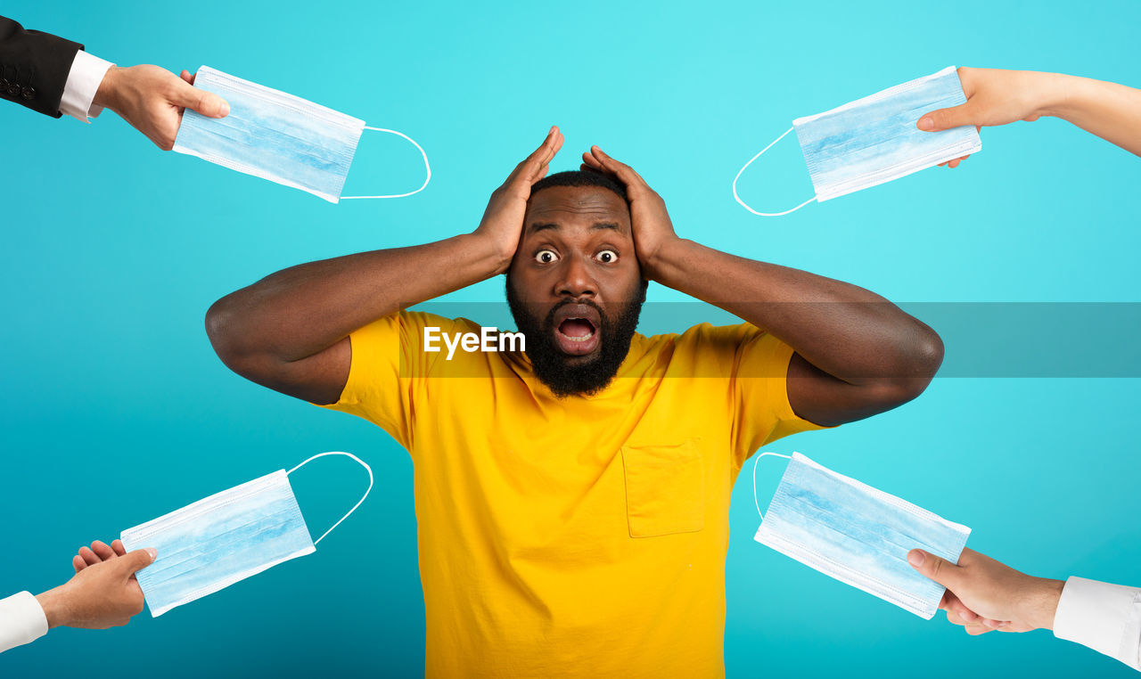 Cropped hands holding masks by shocked man against blue background