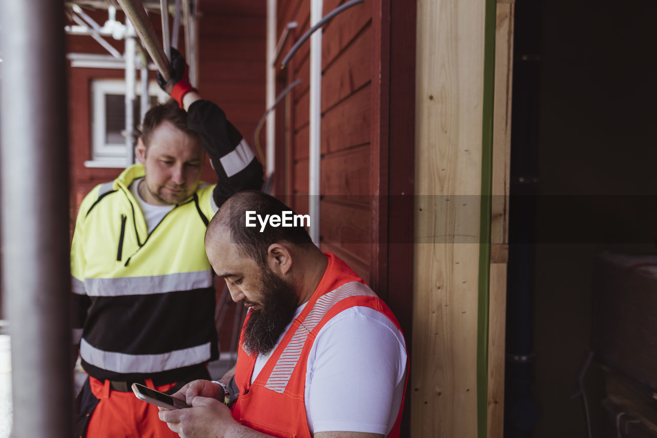 Male construction worker looking at bearded colleague using smart phone at site