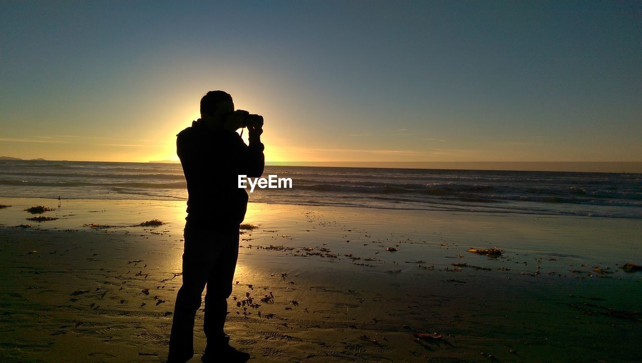 Full length of rear view silhouette man photographing sea against sky at beach