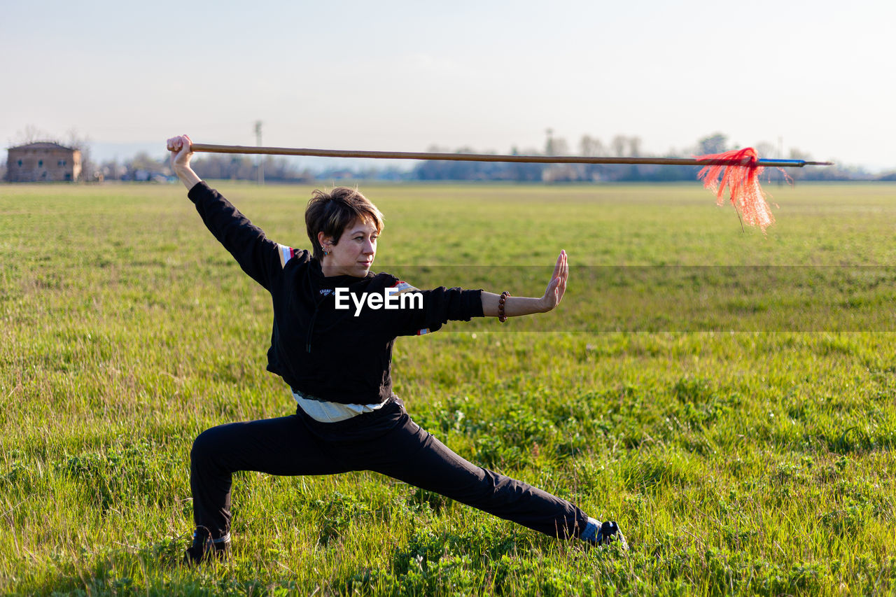 Caucasian young woman practicing wushu martial art on a green meadow with a long spear