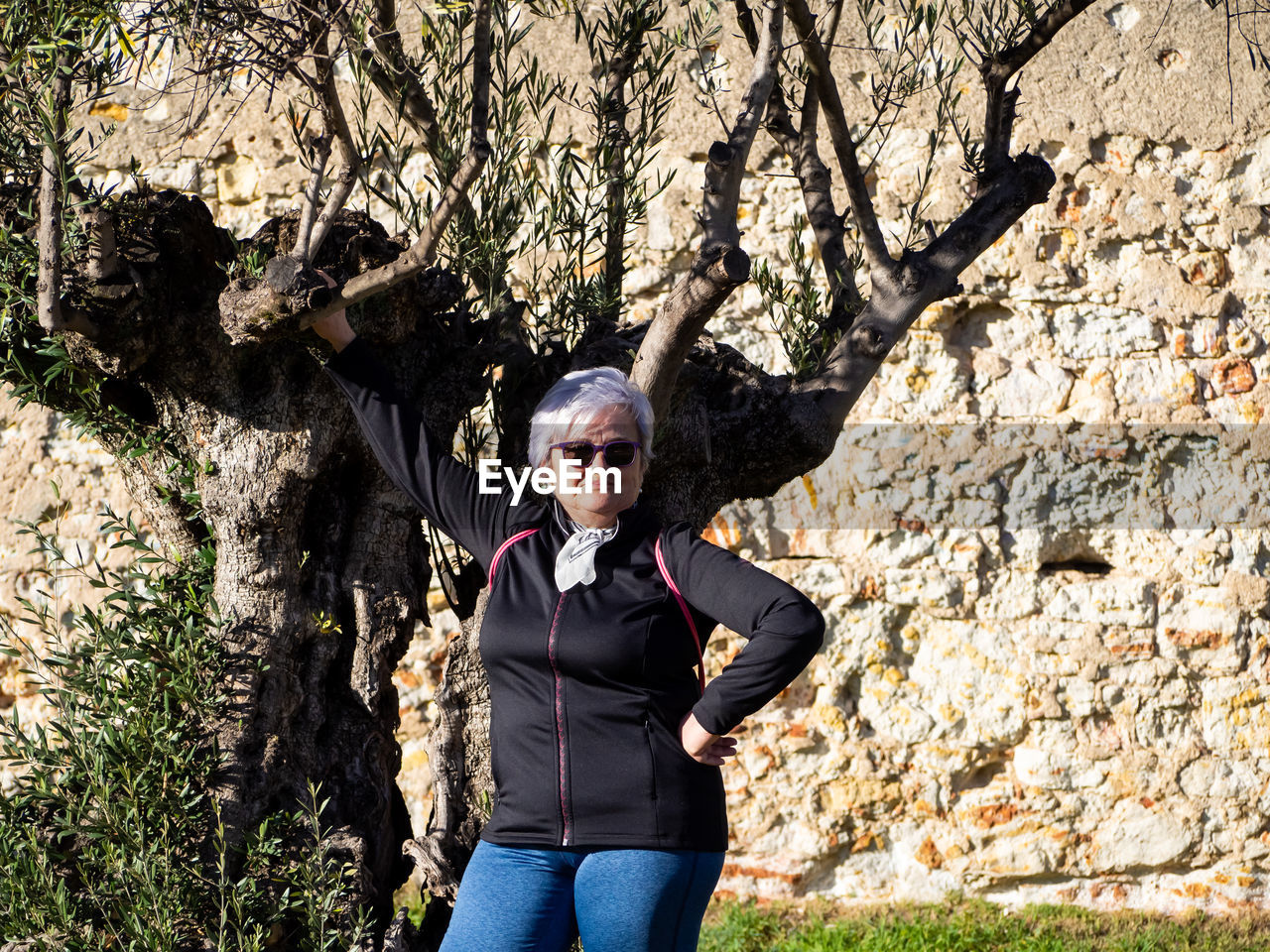 Portrait of senior woman standing by tree during sunny day