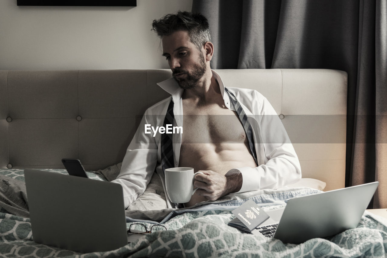 Fit muscular businessman in bed with computers drinking coffee.
