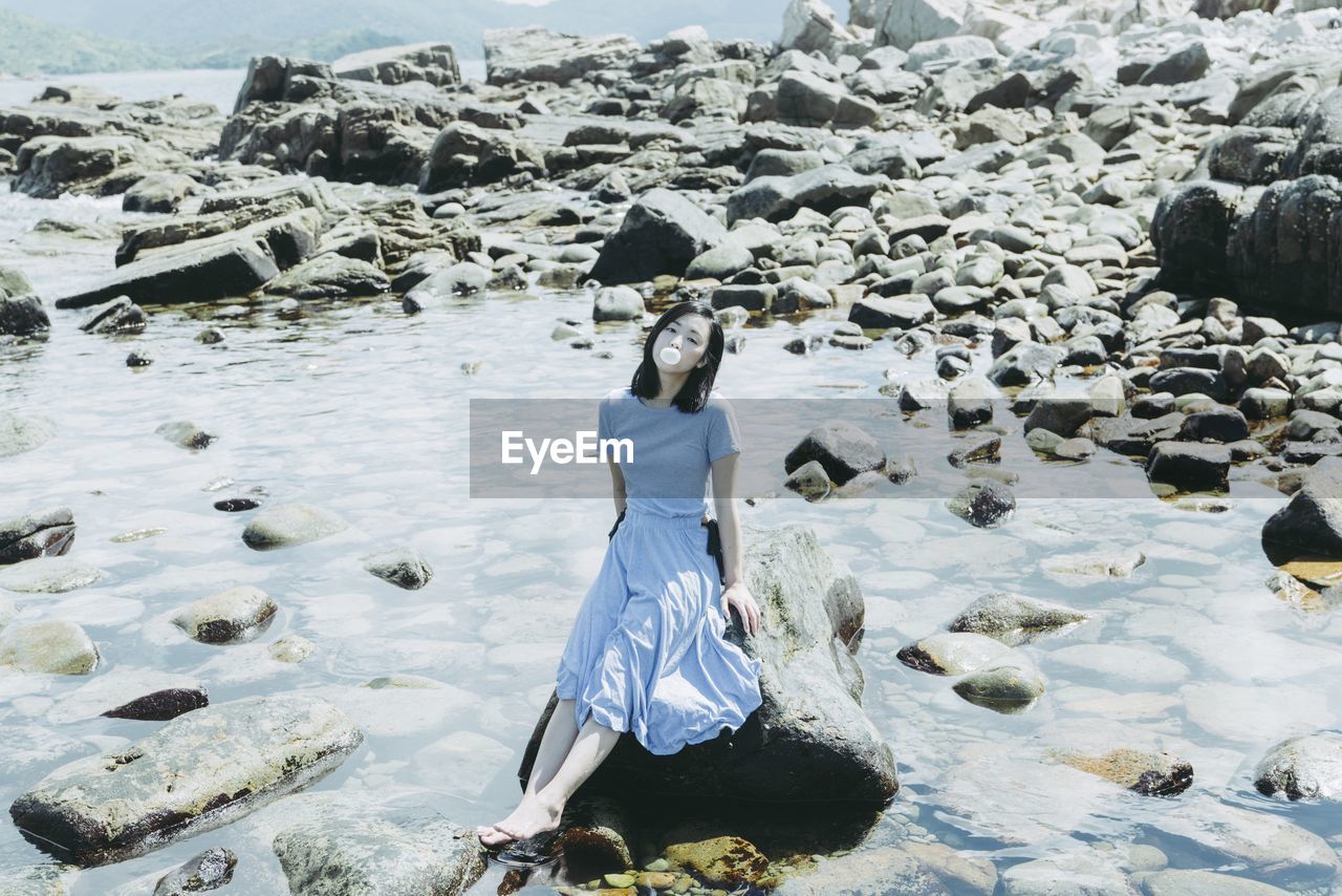 High angle view of beautiful woman blowing bubble gum while sitting on rock at beach