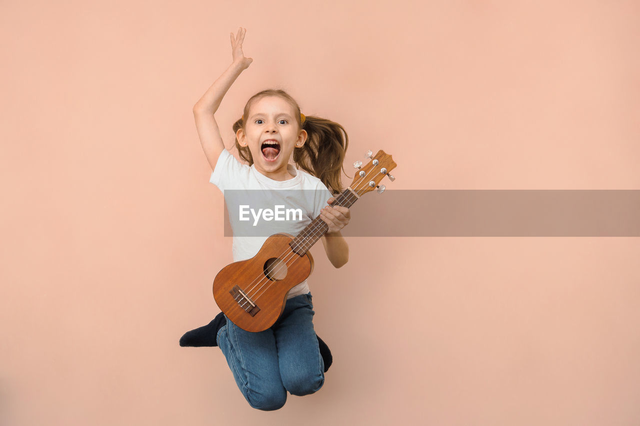 music, musical instrument, one person, string instrument, arts culture and entertainment, musician, happiness, studio shot, copy space, emotion, performance, smiling, portrait, guitar, indoors, cheerful, child, women, vitality, childhood, enjoyment, colored background, musical equipment, fun, full length, mouth open, blond hair, front view, positive emotion, person, looking at camera, adult, holding, human mouth, joy, excitement, female, skill, casual clothing, shouting, lifestyles