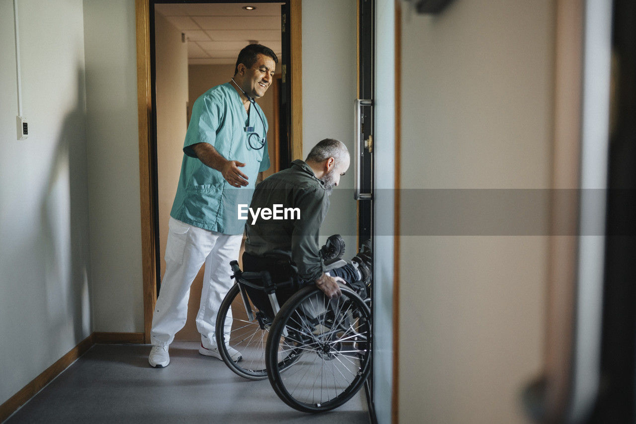Happy doctor assisting patient in wheelchair at corridor of clinic