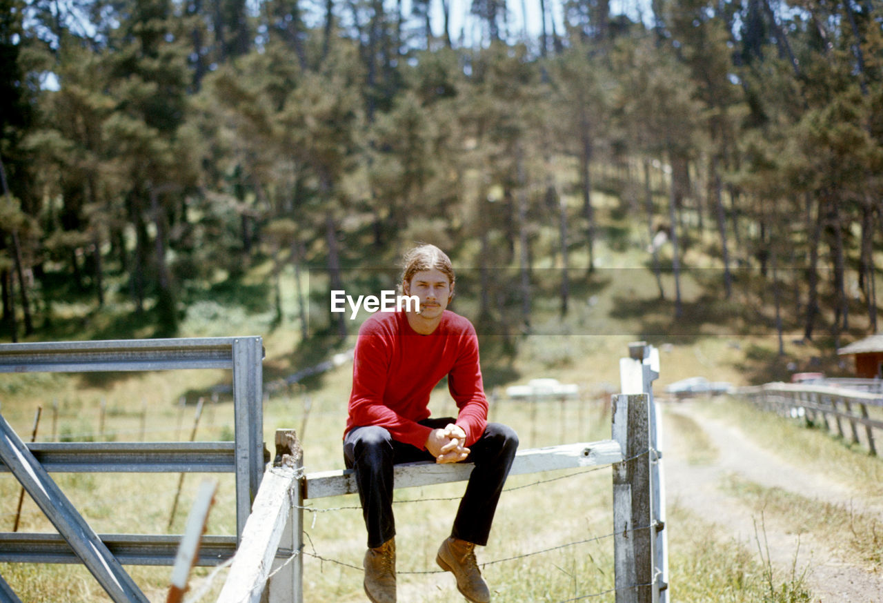 Portrait of young man sitting on fence against trees