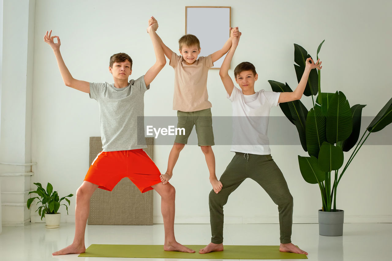 A happy boy and two teenagers perform group yoga exercises with support while sitting on a  mat.